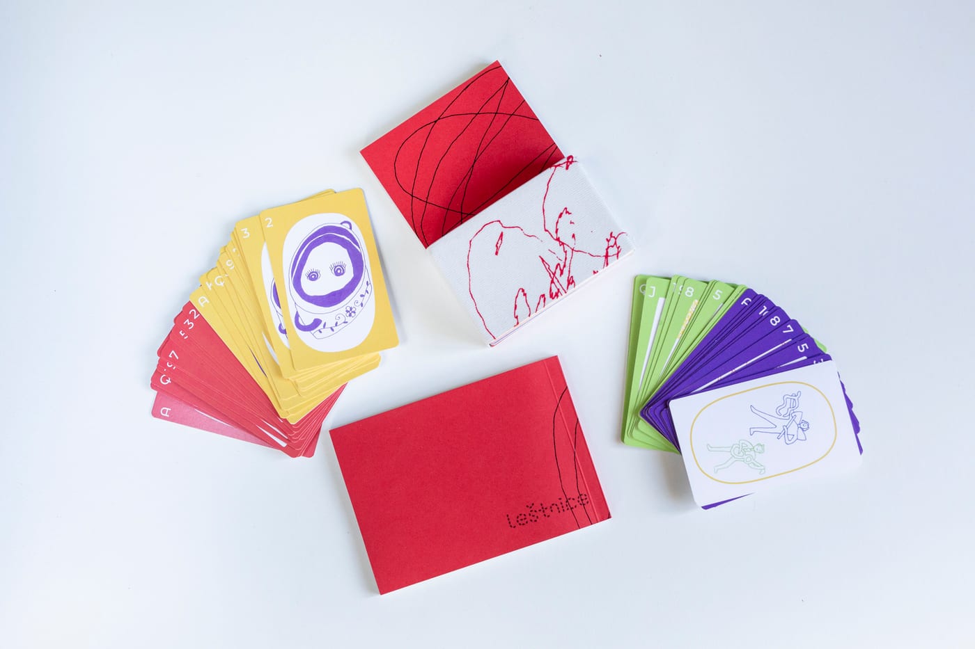 From above on white: fanned red and yellow cards, a red booklet, the slipcase with red drawing, and fanned green and purple cards.