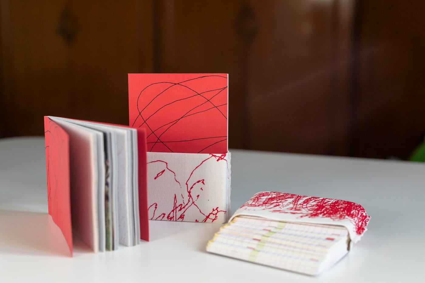 The set on a white table: the red book standing open, a card in the white slipcase printed with red drawing, and a deck in a paper band.