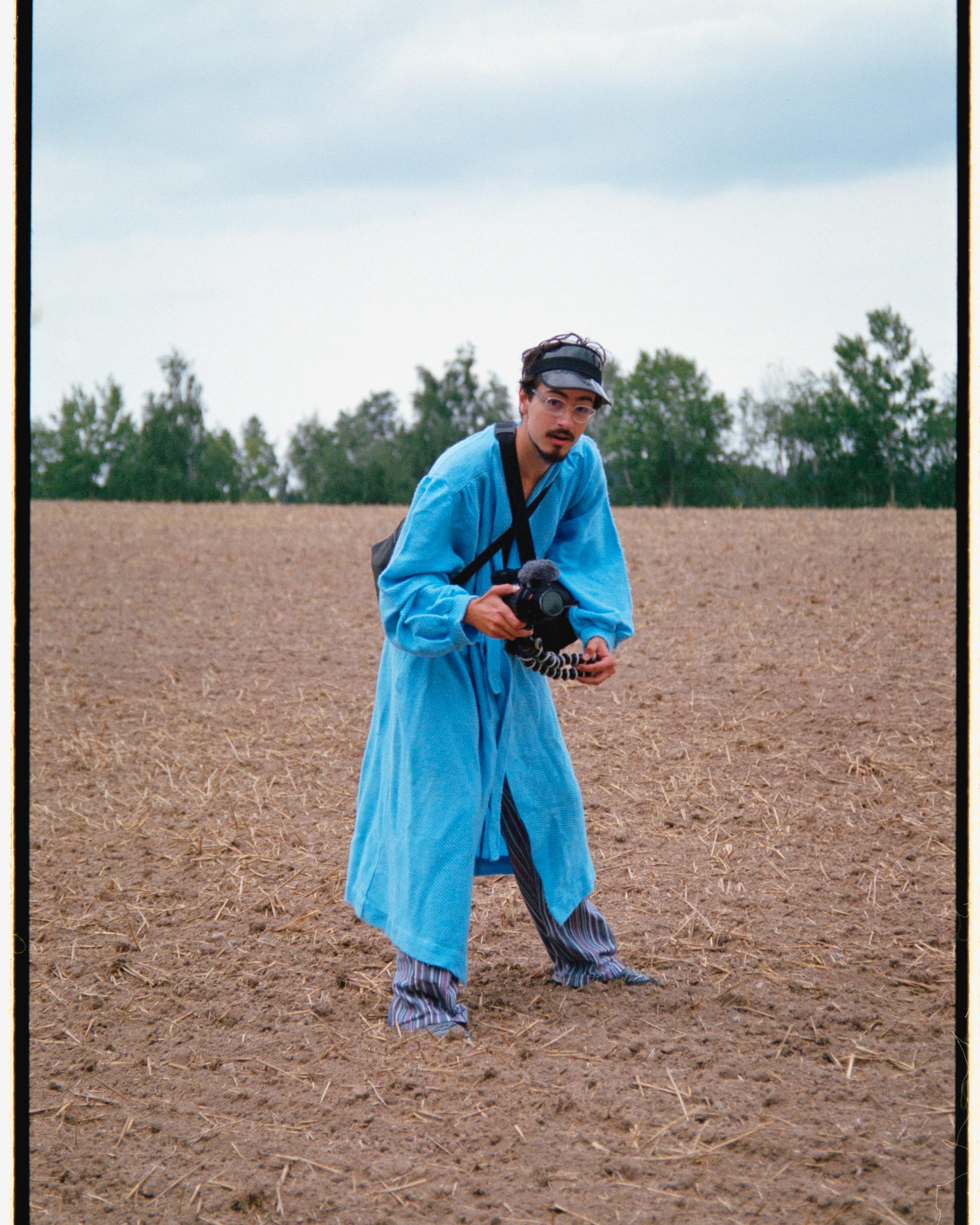 In colour: a man in a long blue robe and a cap stands in a ploughed field, holding a camera at his waist.