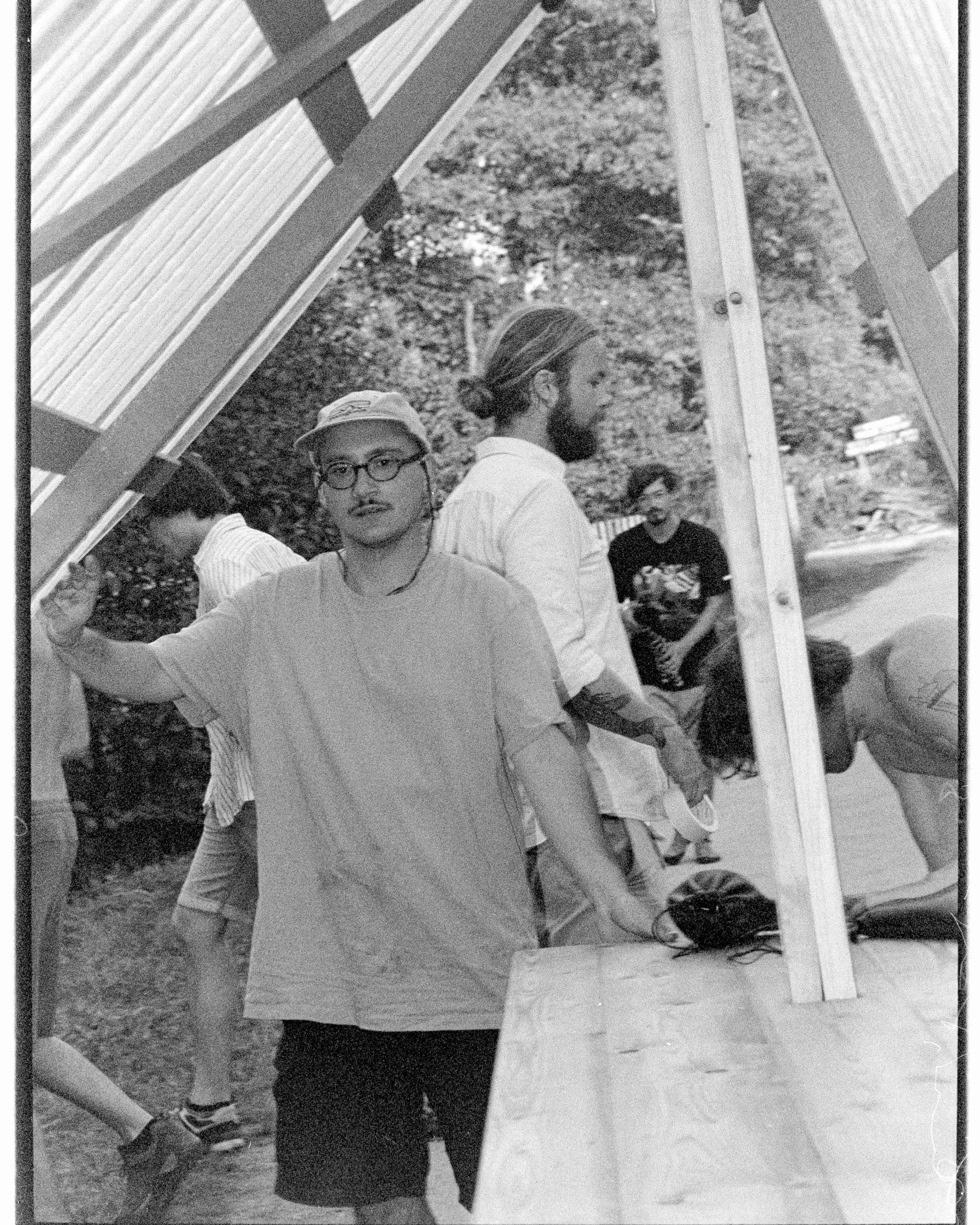 Black and white: a man in a cap and round glasses stands with a hand on a timber frame under a translucent roof, others at work behind him.