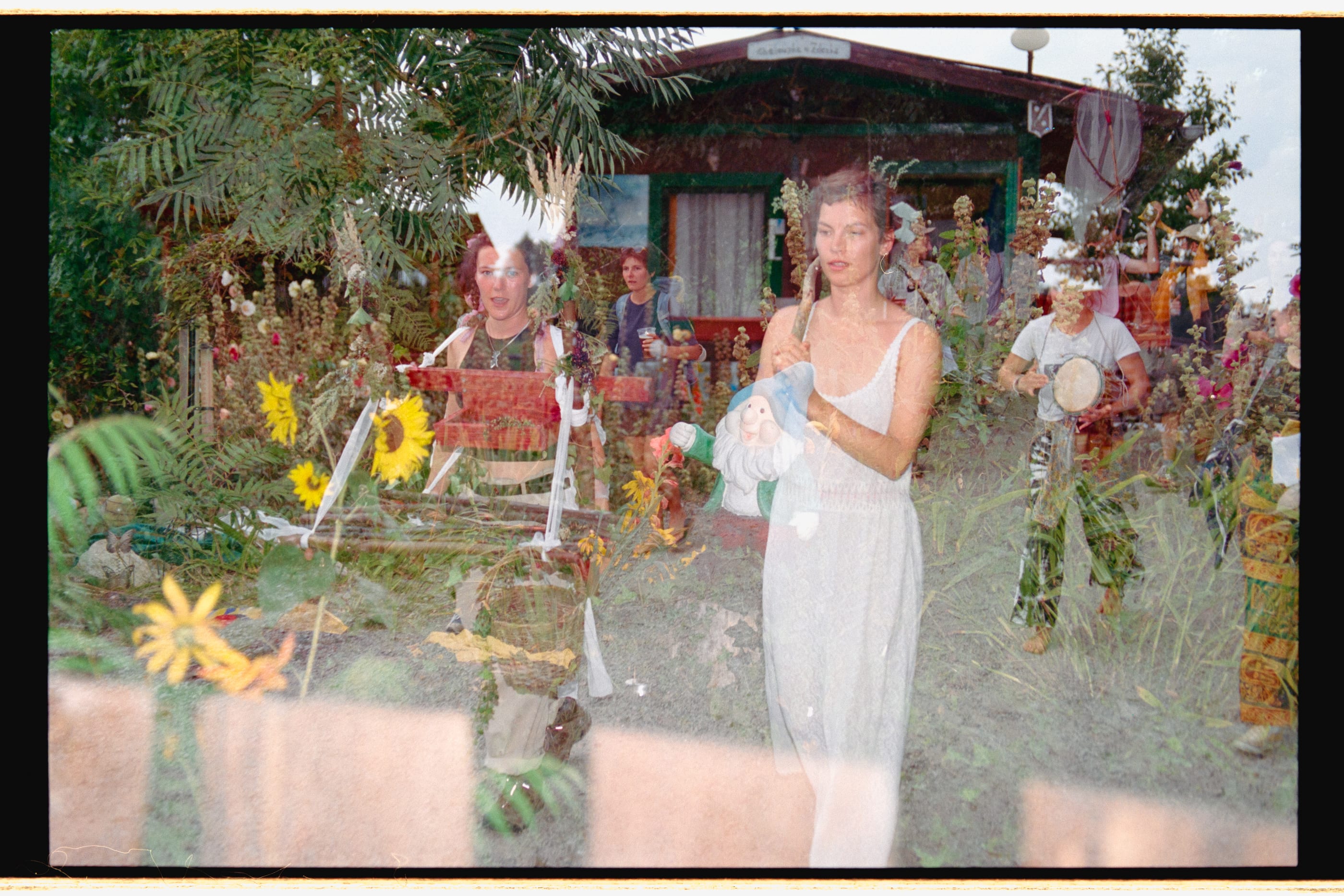 A colour double exposure: a woman in a white dress among sunflowers and garden plants, with musicians and a painted wooden hut behind her.