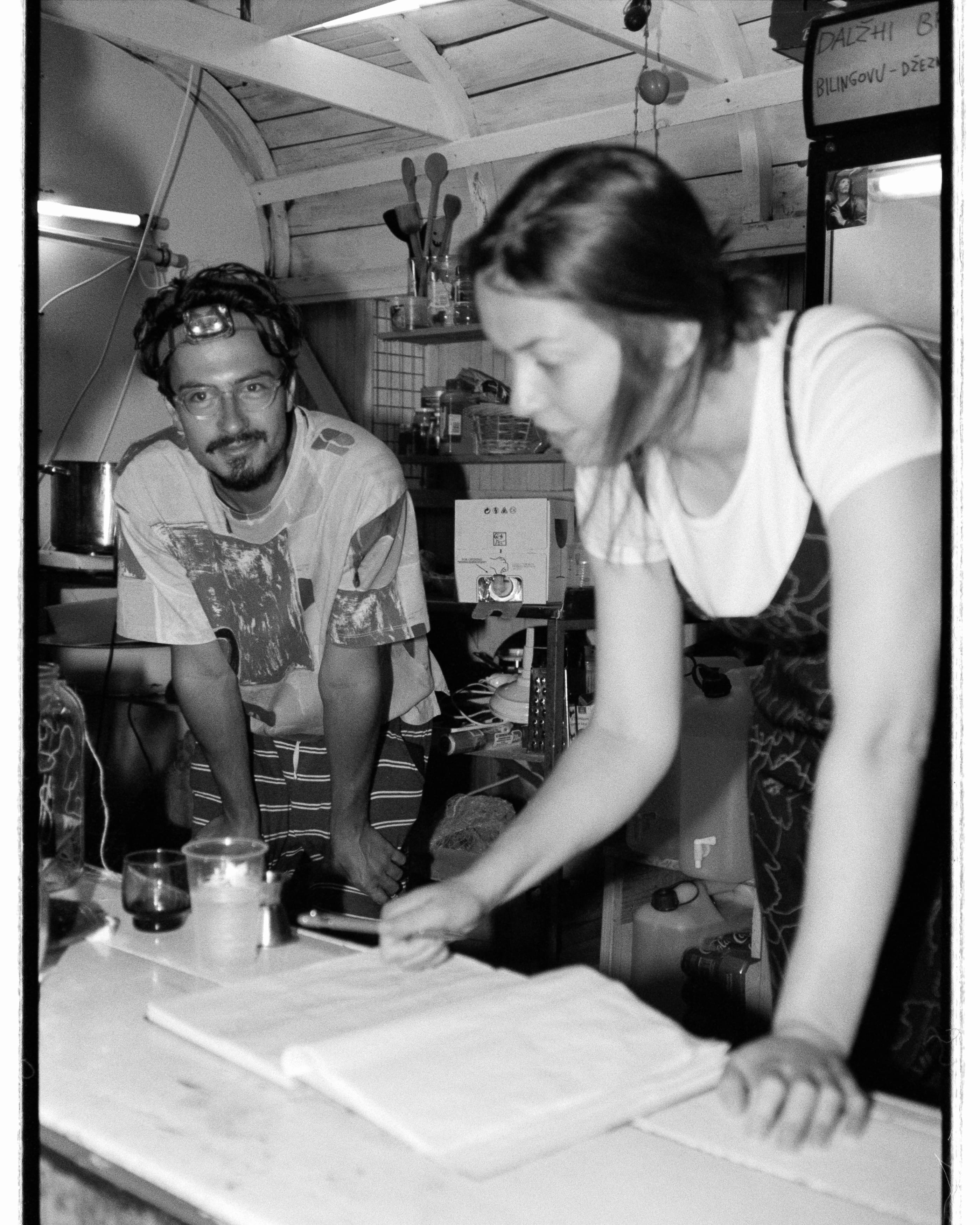 Black and white: in the kitchen shelter a woman leans over an open notebook on the counter while a man with a headlamp looks at the camera.