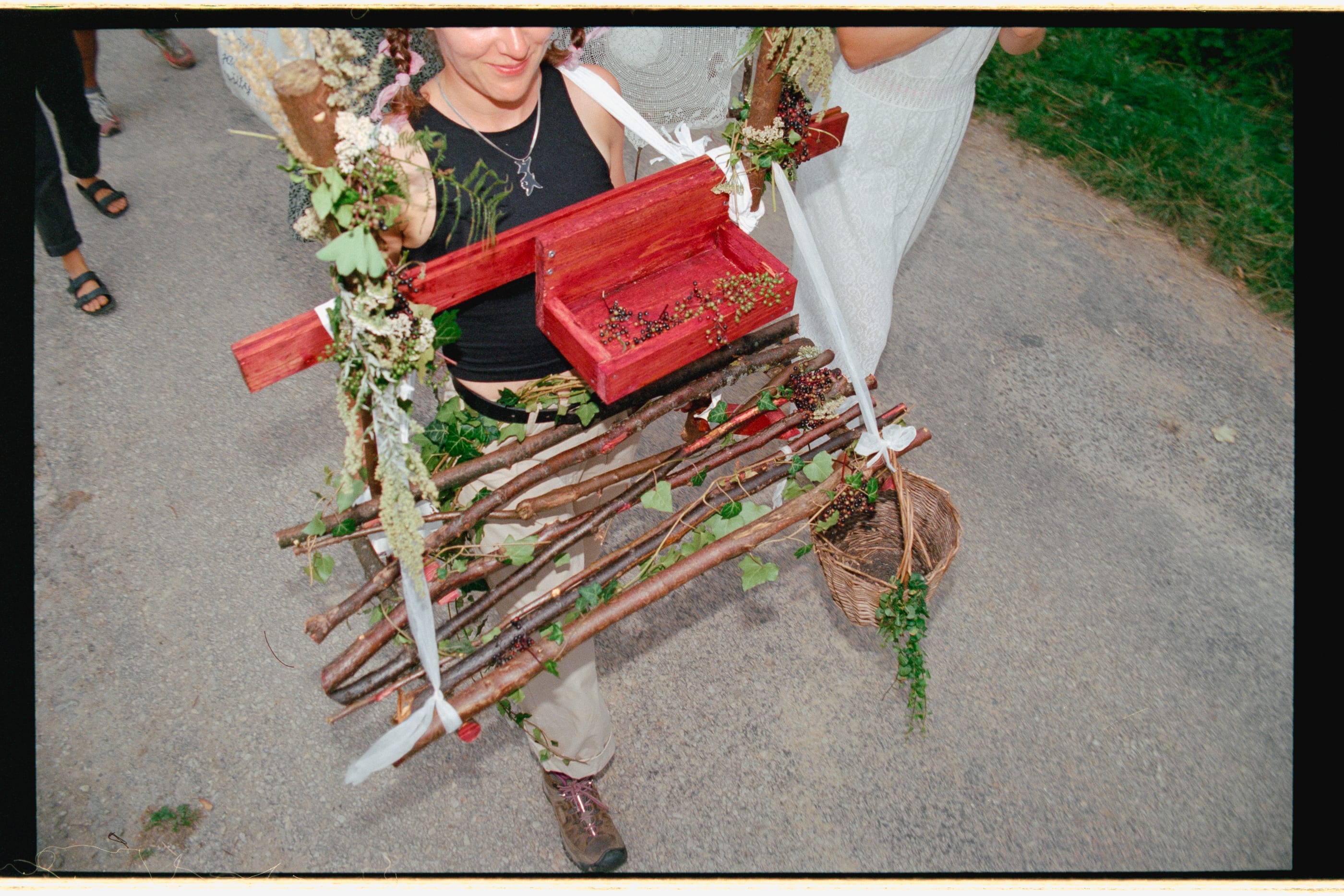 In colour, from above: a woman carries a frame of woven branches hung with ribbons, leaves and a red box along a road.