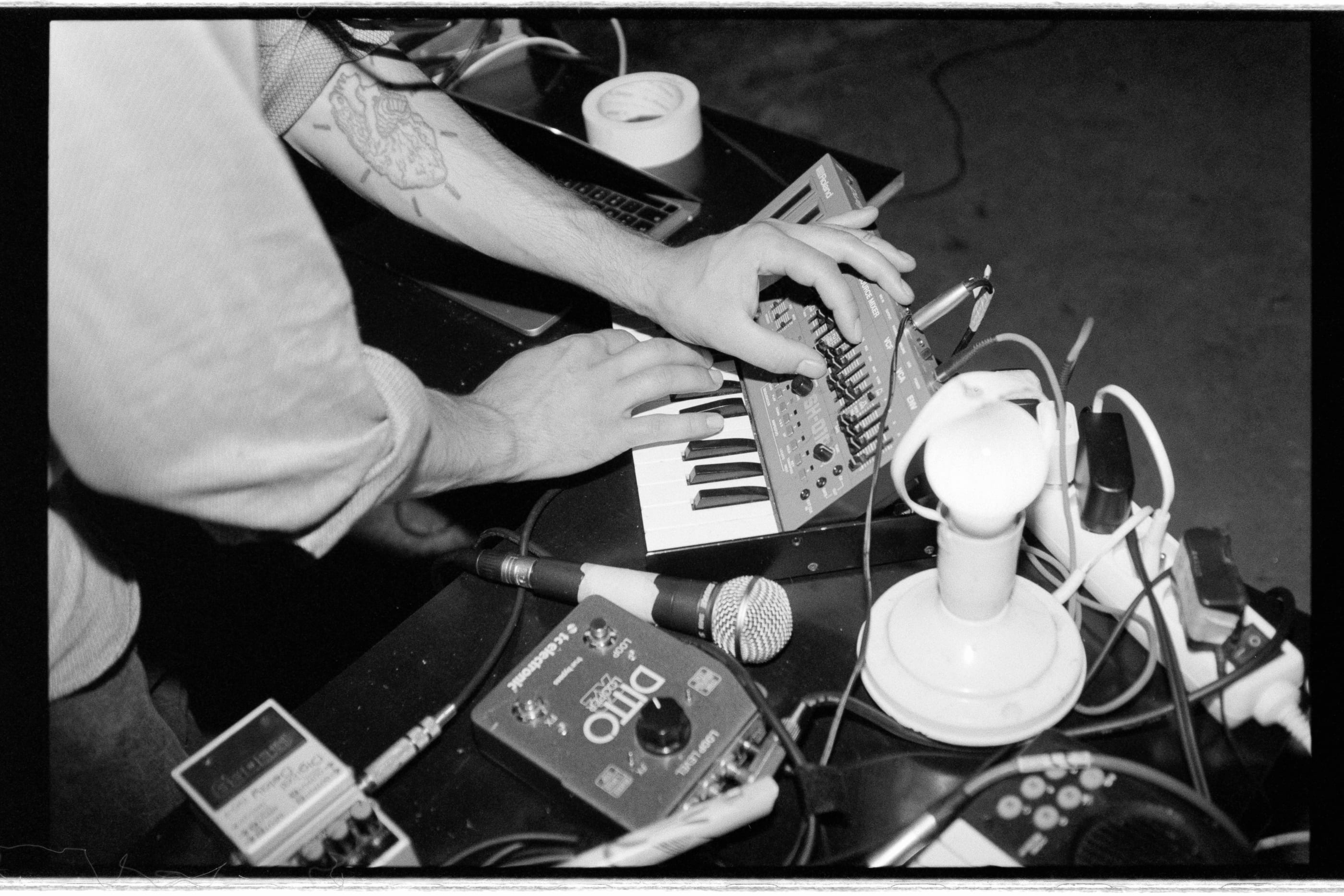 Black and white, from above: tattooed hands play a small synthesizer on a table crowded with effect pedals, a microphone and cables.