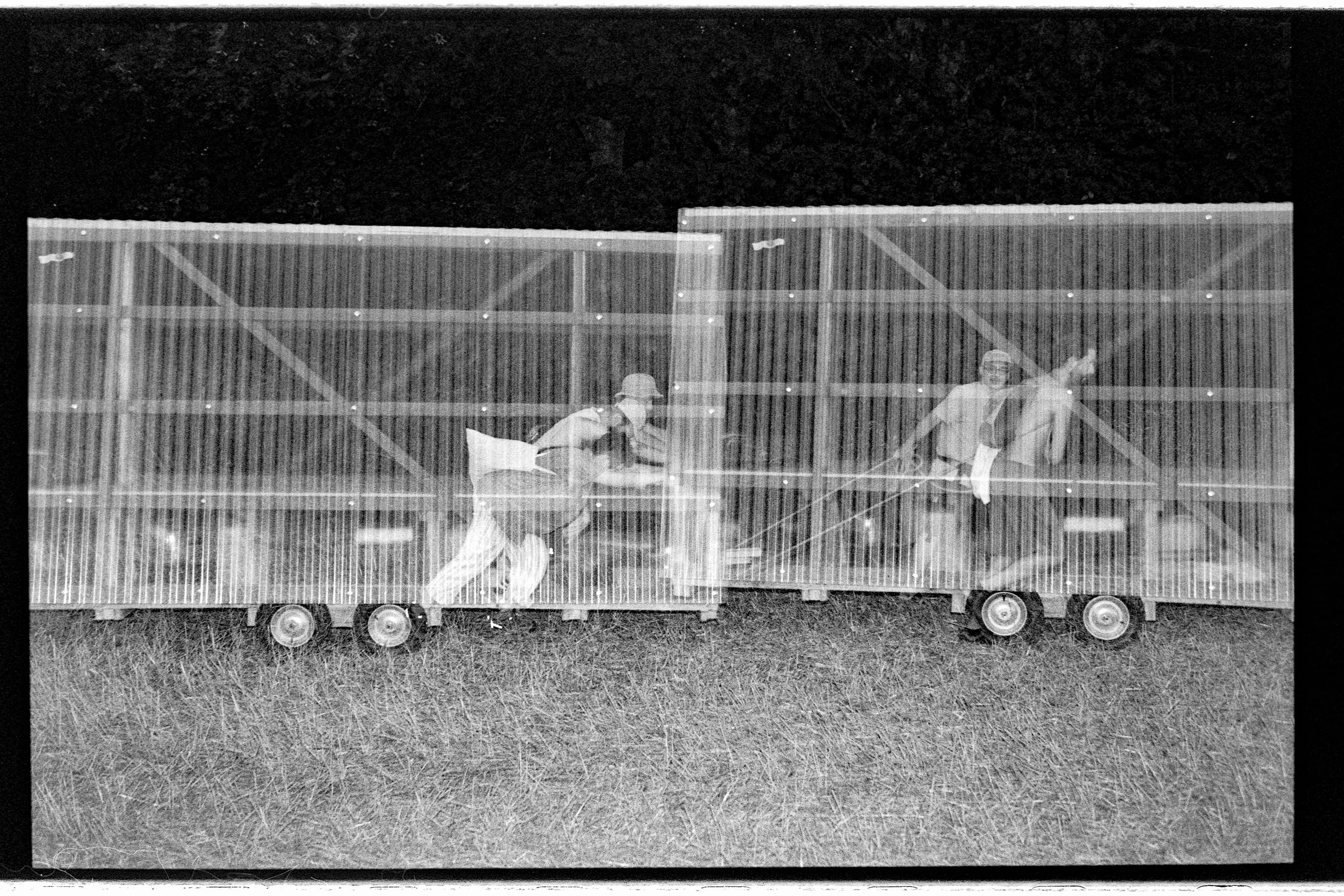 Printed as a negative: two people push two wheeled walls of translucent sheeting across a field.