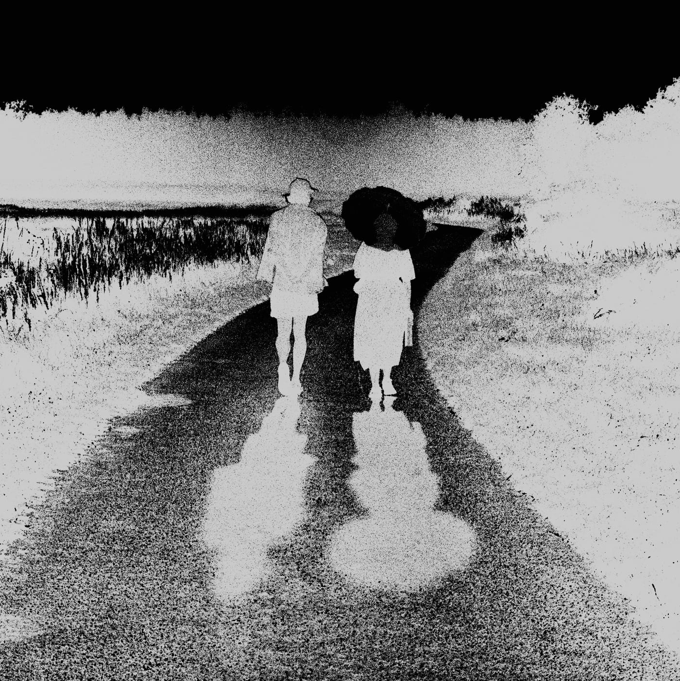 Two people walk down a wet lane, one under a parasol, printed as a negative so the road is dark and the sky black.