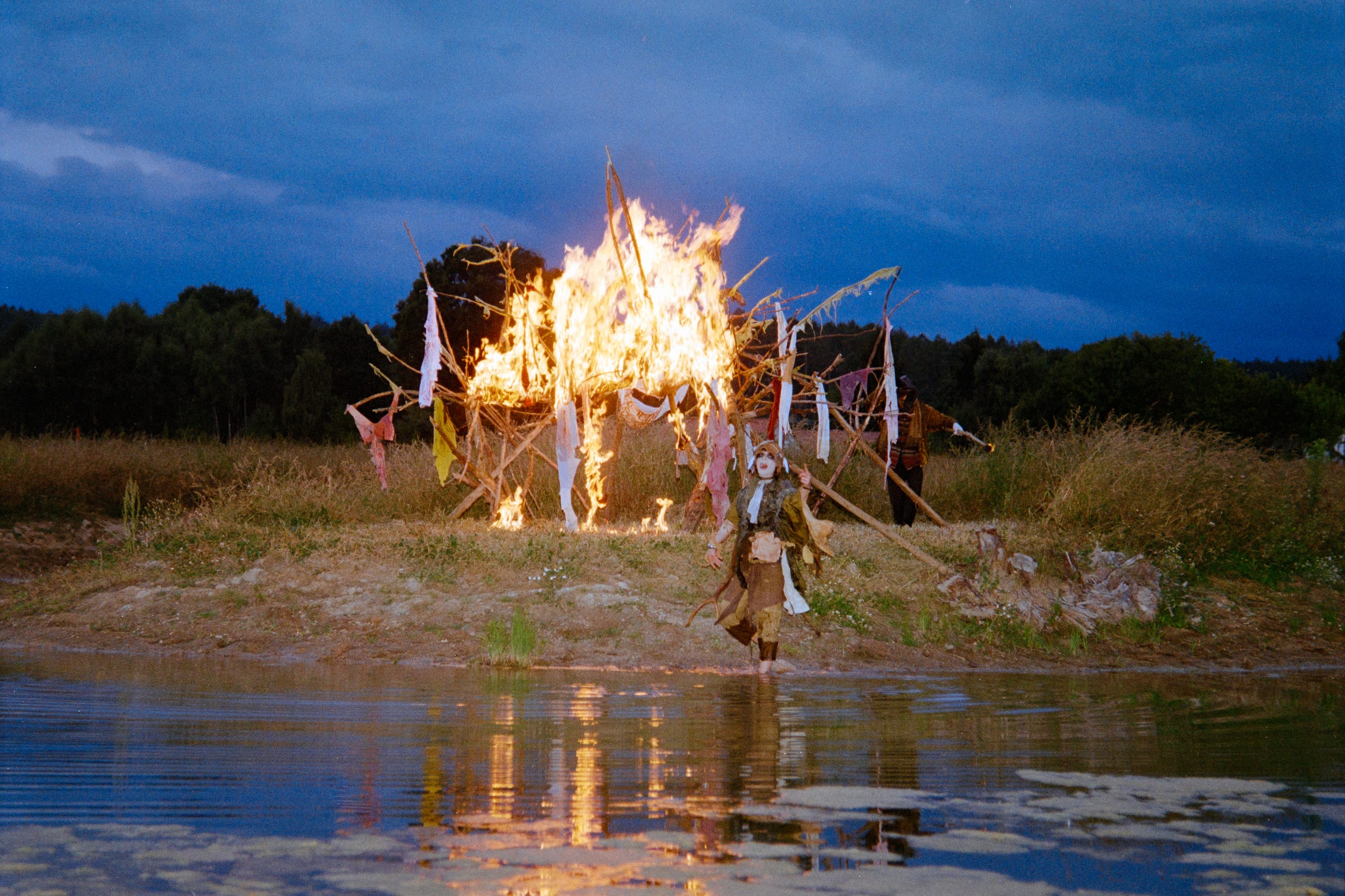 At dusk a wooden structure hung with strips of fabric burns at the edge of a pond, reflected in the water, with a costumed figure beside it.
