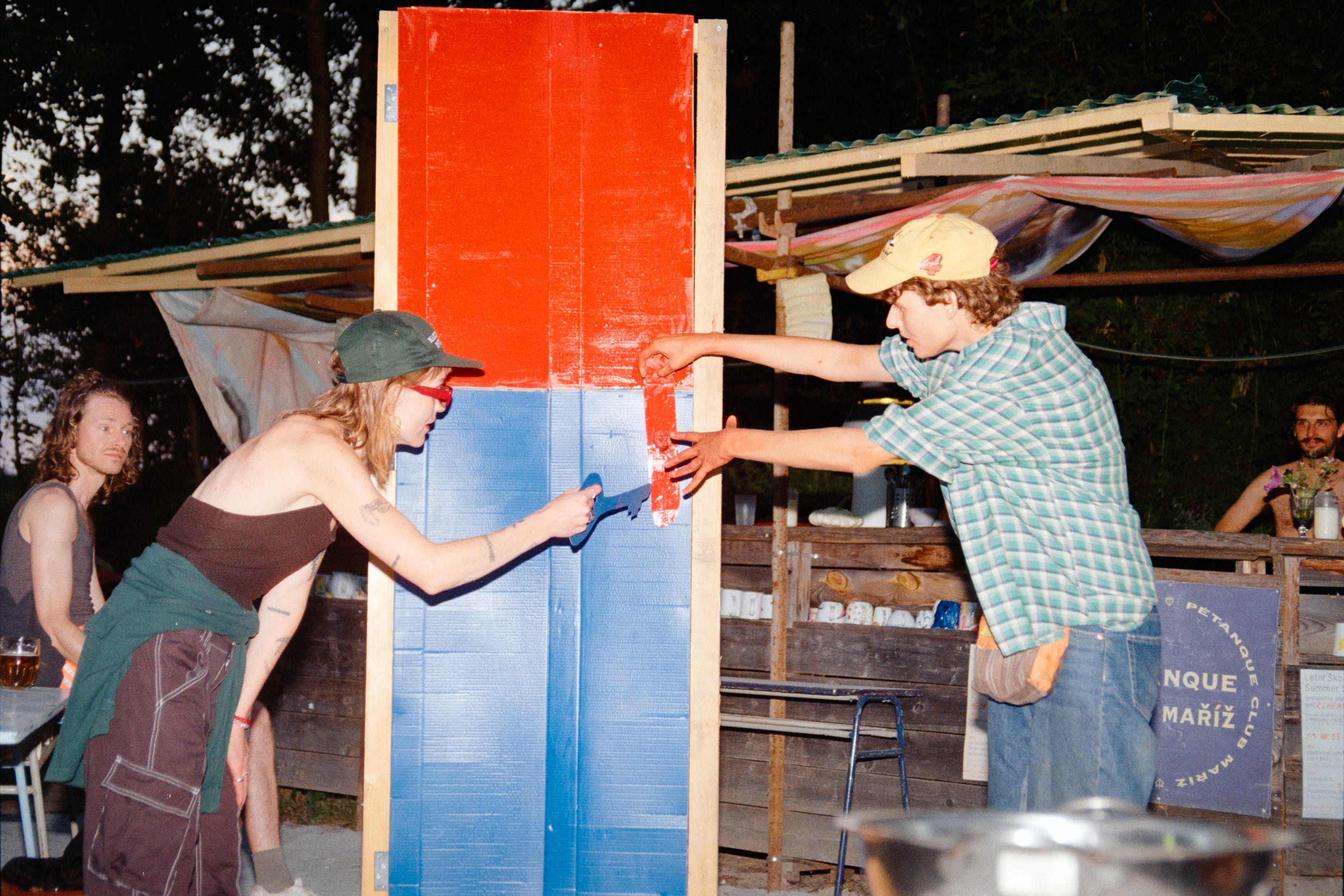 Two people paint a tall red and blue wooden panel at dusk in front of the Maringotka kitchen shelter.