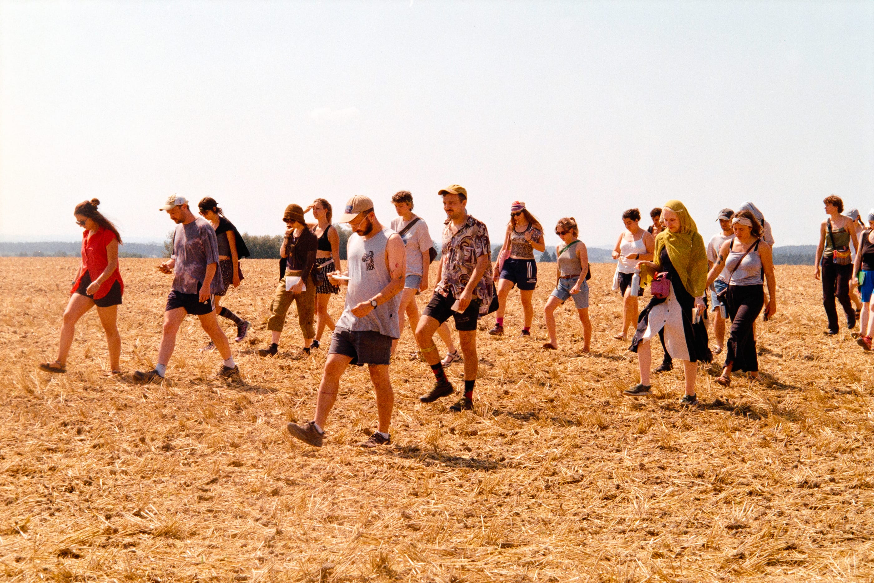 A long line of about twenty people walks across a harvested, golden stubble field under a pale sky.