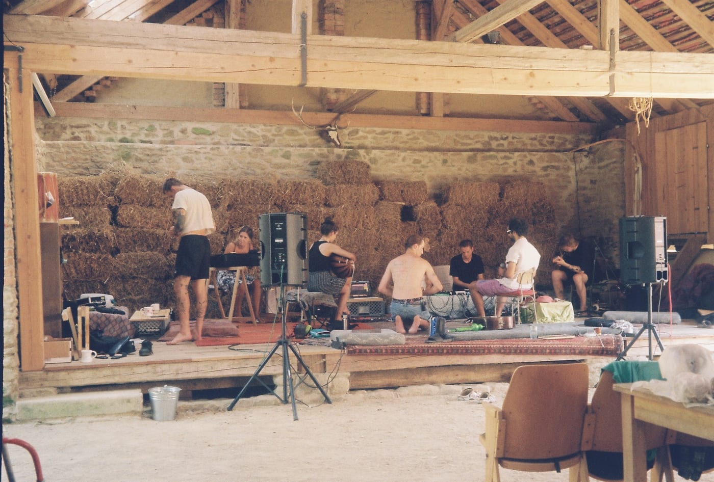 A wide barn with a stage of hay bales. People sit and kneel on rugs among loudspeakers and cables, playing and recording.