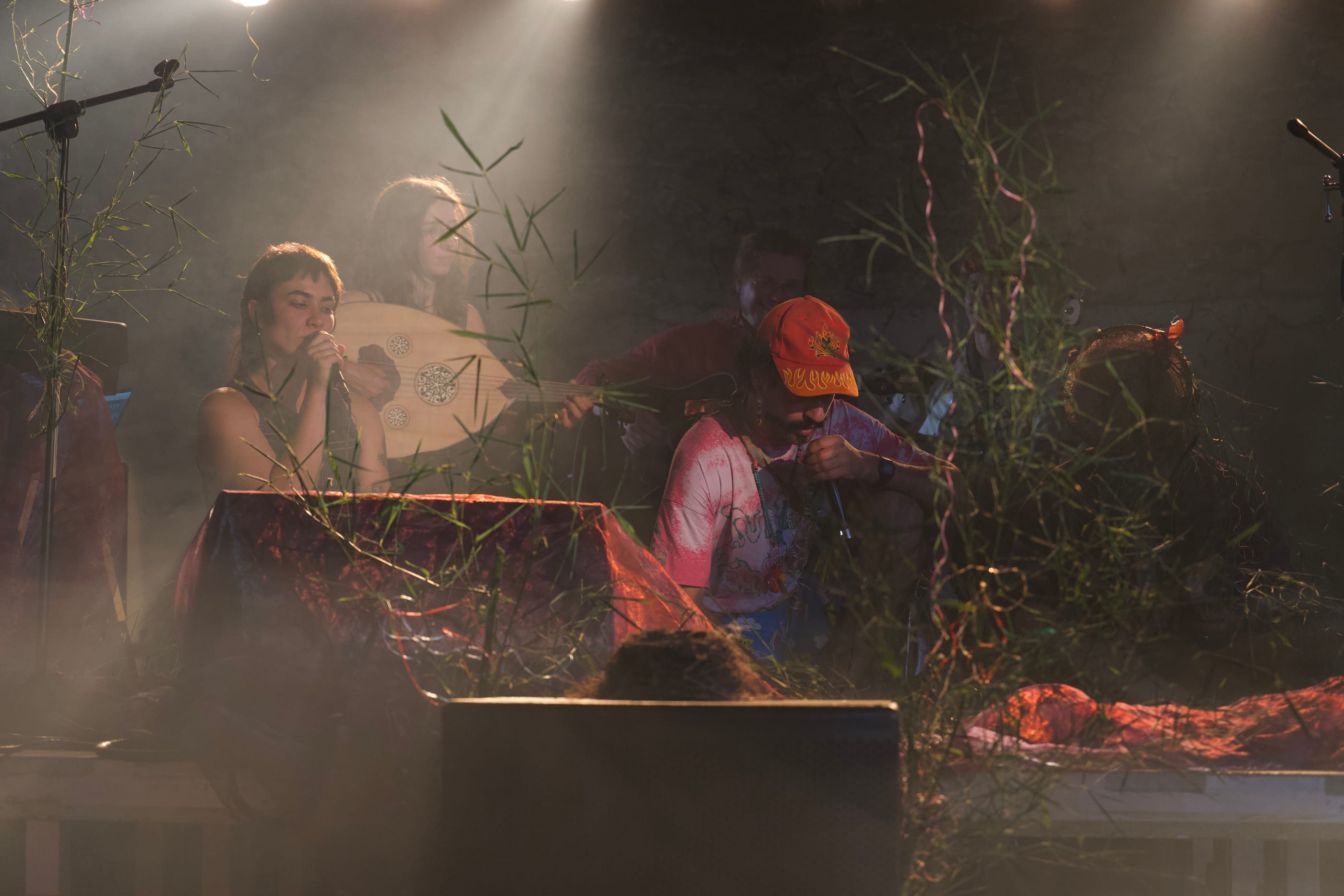 A concert on a stage behind grasses and paper streamers: a woman at a microphone, another with an oud, a man in an orange cap singing.