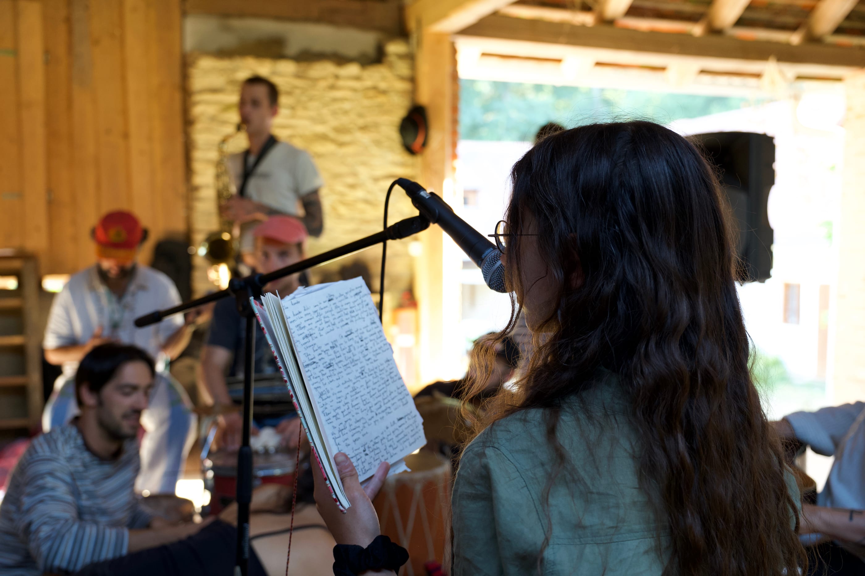 In the barn, a woman with long dark hair reads from a handwritten notebook into a microphone. Behind her a saxophonist and two drummers in caps.