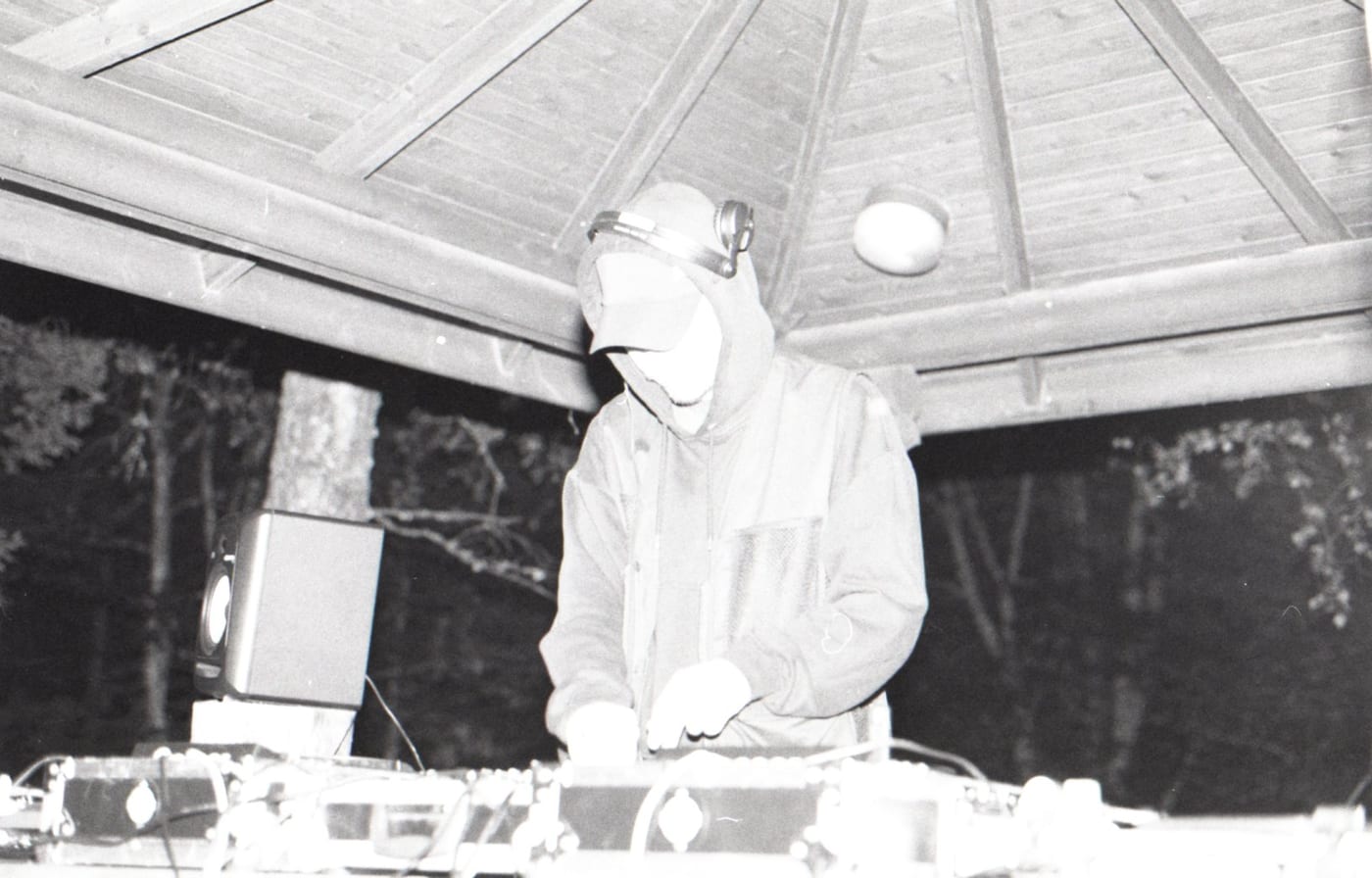 Printed as a negative: a person in a cap and headphones plays records under the wooden roof of a pavilion at night.