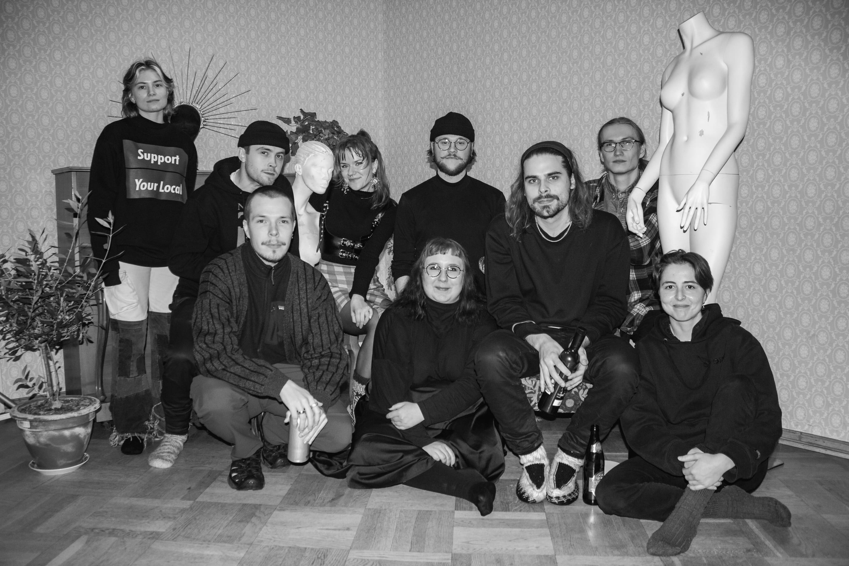 Nine people sitting and standing together in a wallpapered room beside a mannequin, in black and white.