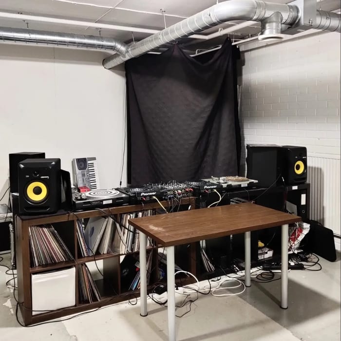 Turntables, a mixer and two speakers on a shelf of records, with a keyboard behind them and a black cloth on the wall.