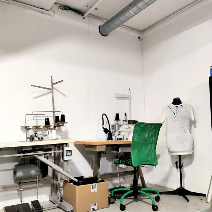 Two industrial sewing machines on work tables, a green office chair and a dress form in a white garment, in a white room.