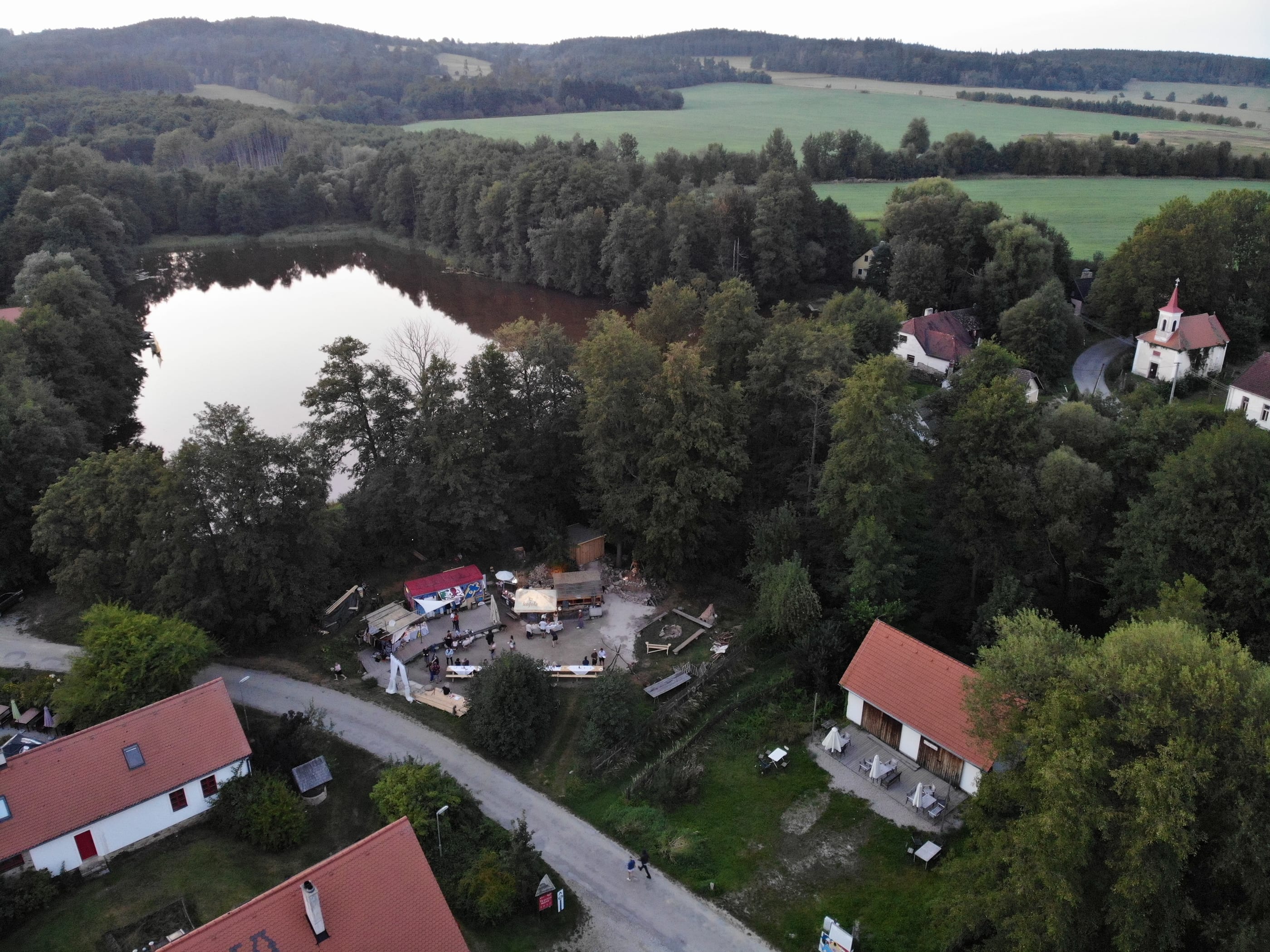 Maříž from above in late summer: a pond, a chapel, a few red roofs among trees, and fields around them.