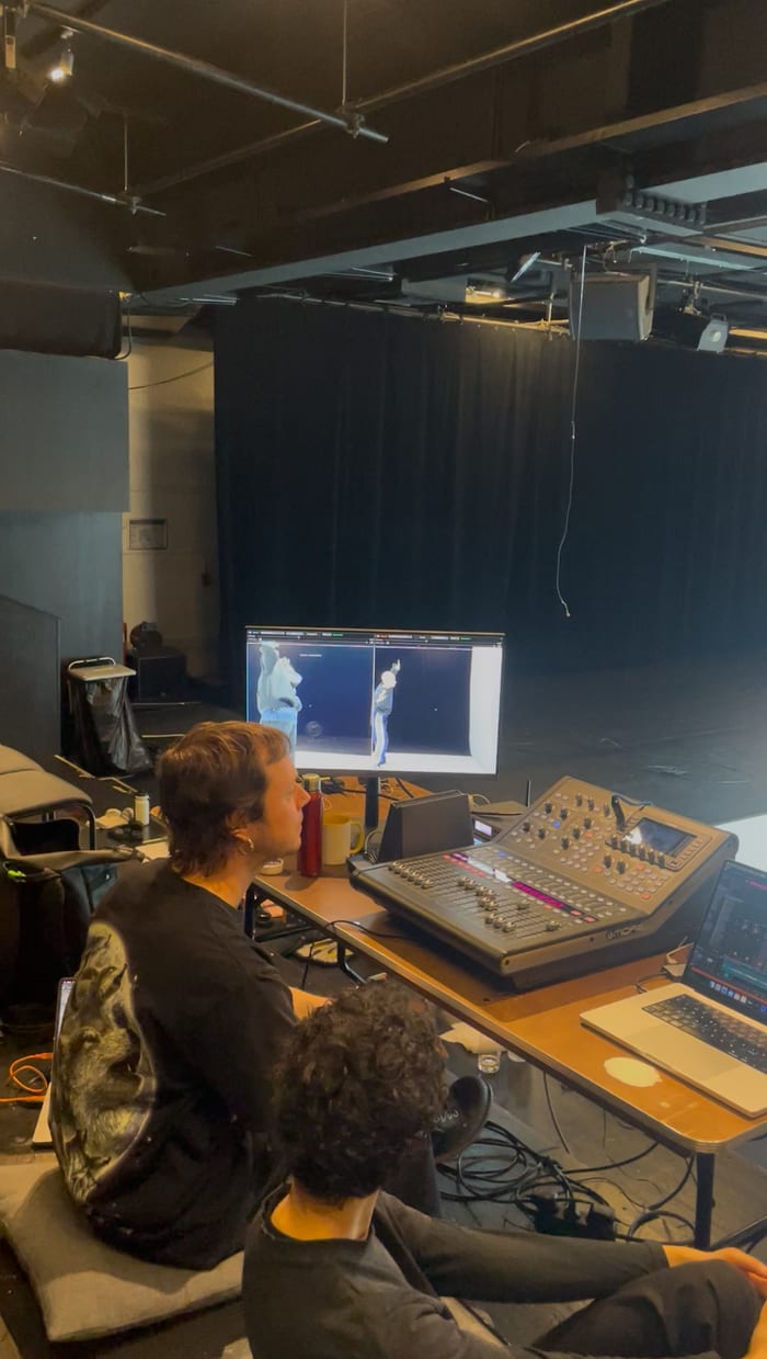 Two people sit on cushions at a low desk in a dark theatre, seen from behind. On the desk a monitor shows the app's two camera pictures with a dancer in each, next to a mixing console and a laptop. The stage curtain is behind.