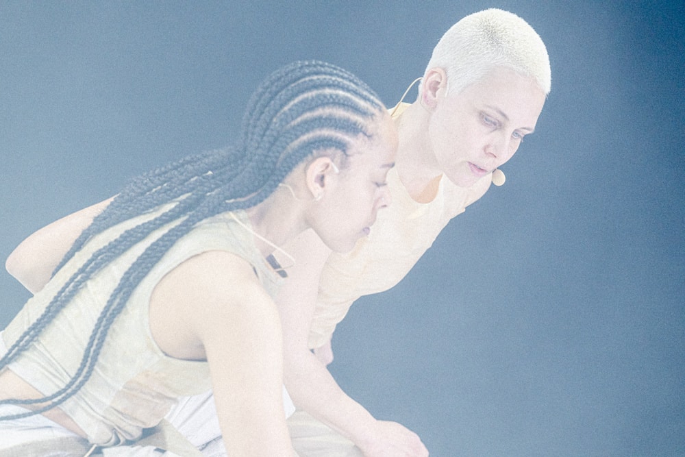 Two dancers bend forward close together, one with long braids and one with short bleached hair, both looking down, in a pale blue haze.