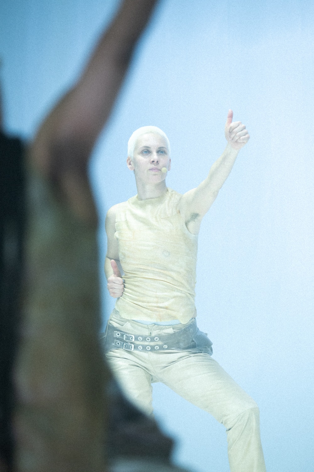 A dancer with short bleached hair and a small microphone at the cheek holds one thumb up high and one at the hip, in pale light. Another dancer's arm is a blur in the foreground.