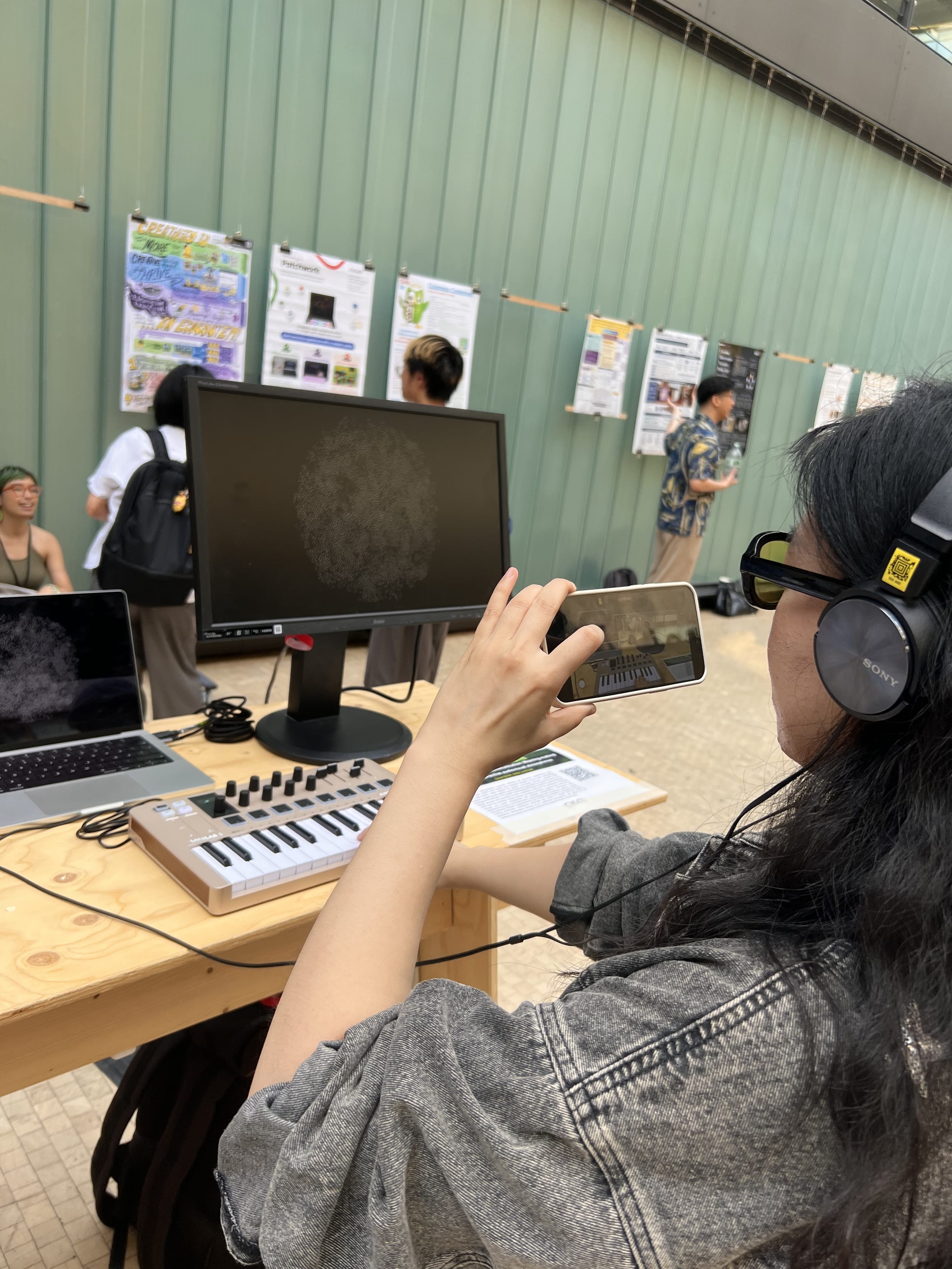 Seen from behind her shoulder, a visitor in headphones and yellow tinted glasses holds up a phone to film the monitor, where a round white swarm fills the screen. The MIDI keyboard and a laptop stand on the plywood table, and people look at posters along the green wall in the background.