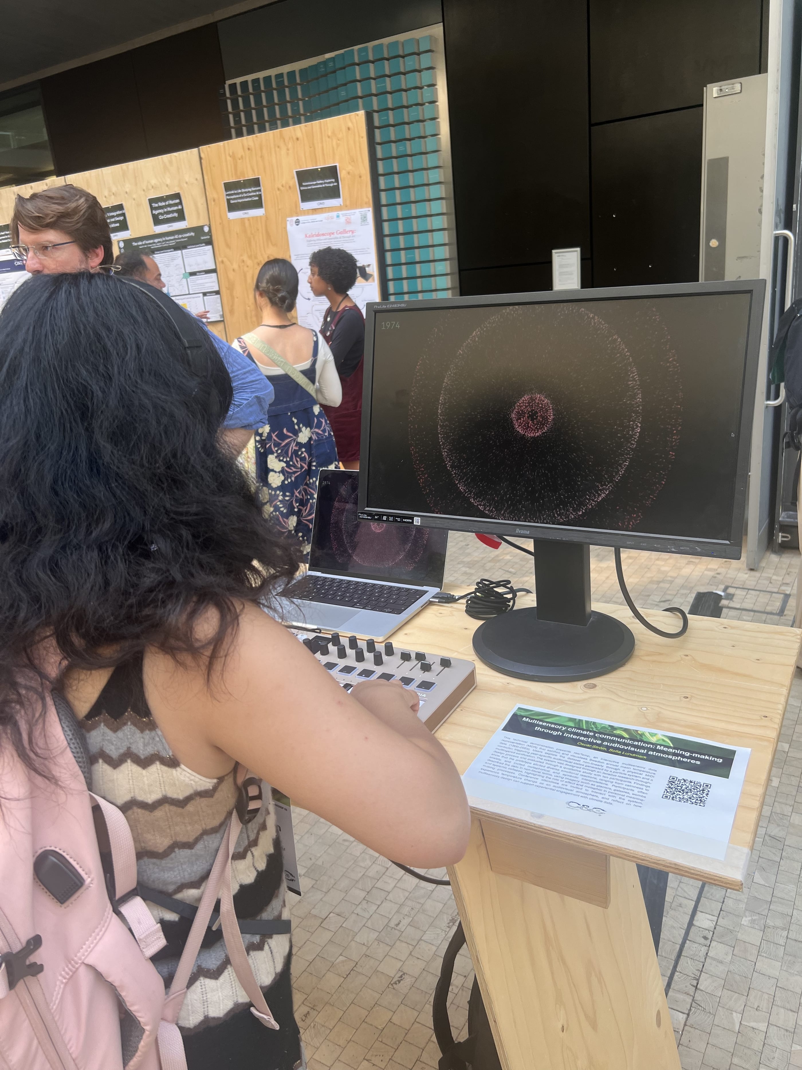 A visitor with long dark hair and a pink backpack, seen from behind, plays the MIDI keyboard in front of a monitor showing a red swarm in rings and the year 1974. On a plywood stand beside her lies a printed card with the title Multisensory climate communication and a QR code. Other conference guests talk in the background.